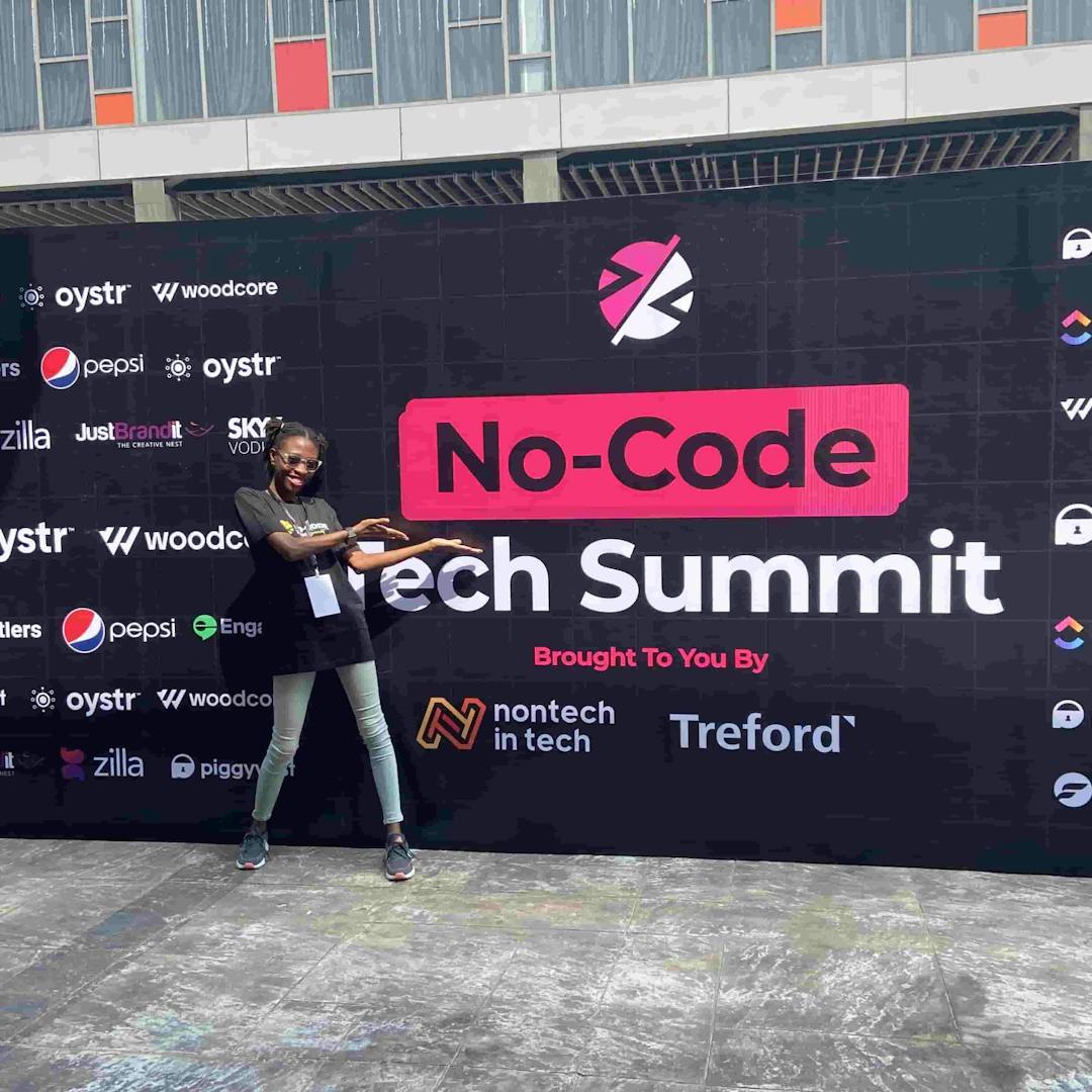 Timonwa is standing in front of a No-code tech summit banner. She is smiling and pointing to the banner.
