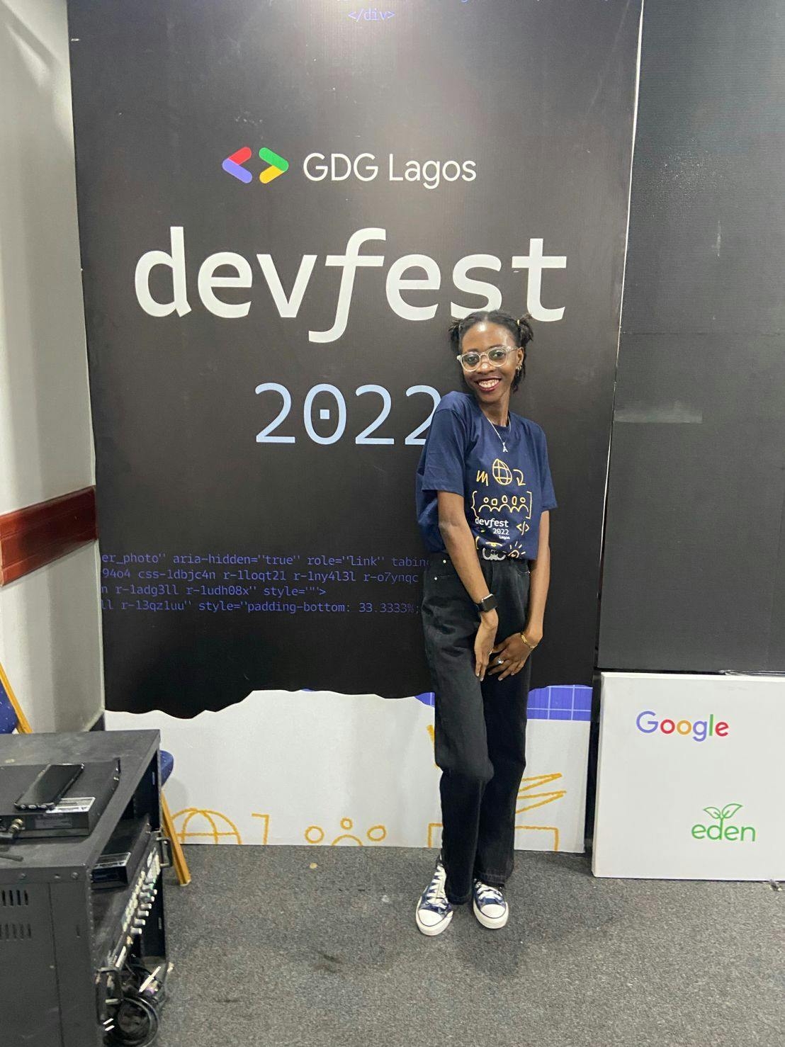 Timonwa is standing and smiling in front of a DevFest 2022 banner. She is wearing DevFest 2022 volunteer t-shirt and a black jeans trousers.